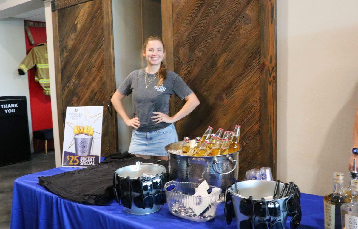 Smiling woman in a gray t-shirt and denim shorts stands behind a table with beer bottles and a promotional sign on display.