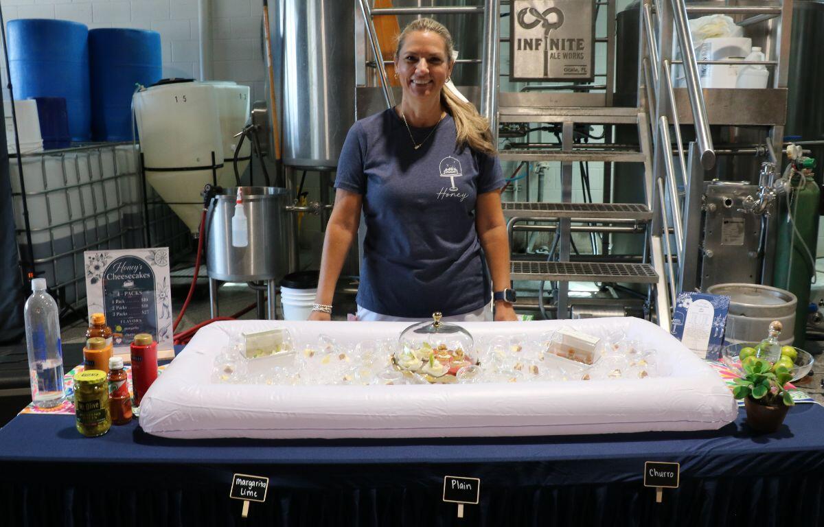 Woman in a navy t‑shirt stands behind a table with an ice-filled display of bite-size cheesecakes under a glass cloche in a brewery setting, with flavor labels nearby (Plain, Margarita Lime, Churro).