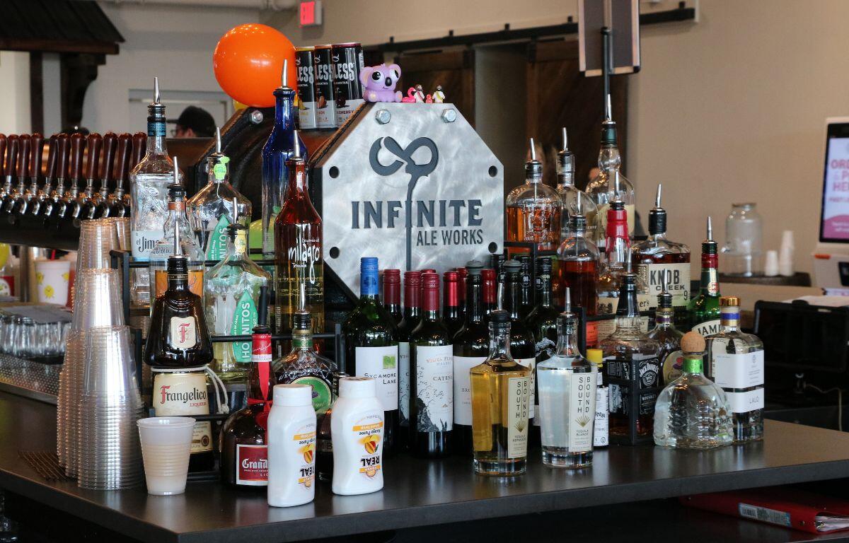 Bar counter filled with assorted liquor bottles and a metal 'Infinite Ale Works' sign at center.