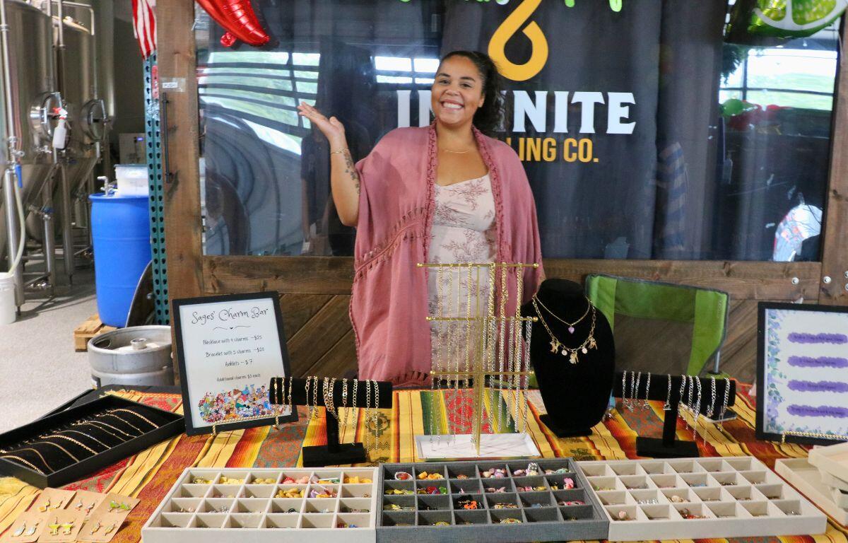 Smiling woman runs a jewelry stall with necklaces on a black bust and bead trays on a colorful striped tablecloth, in a market setting.