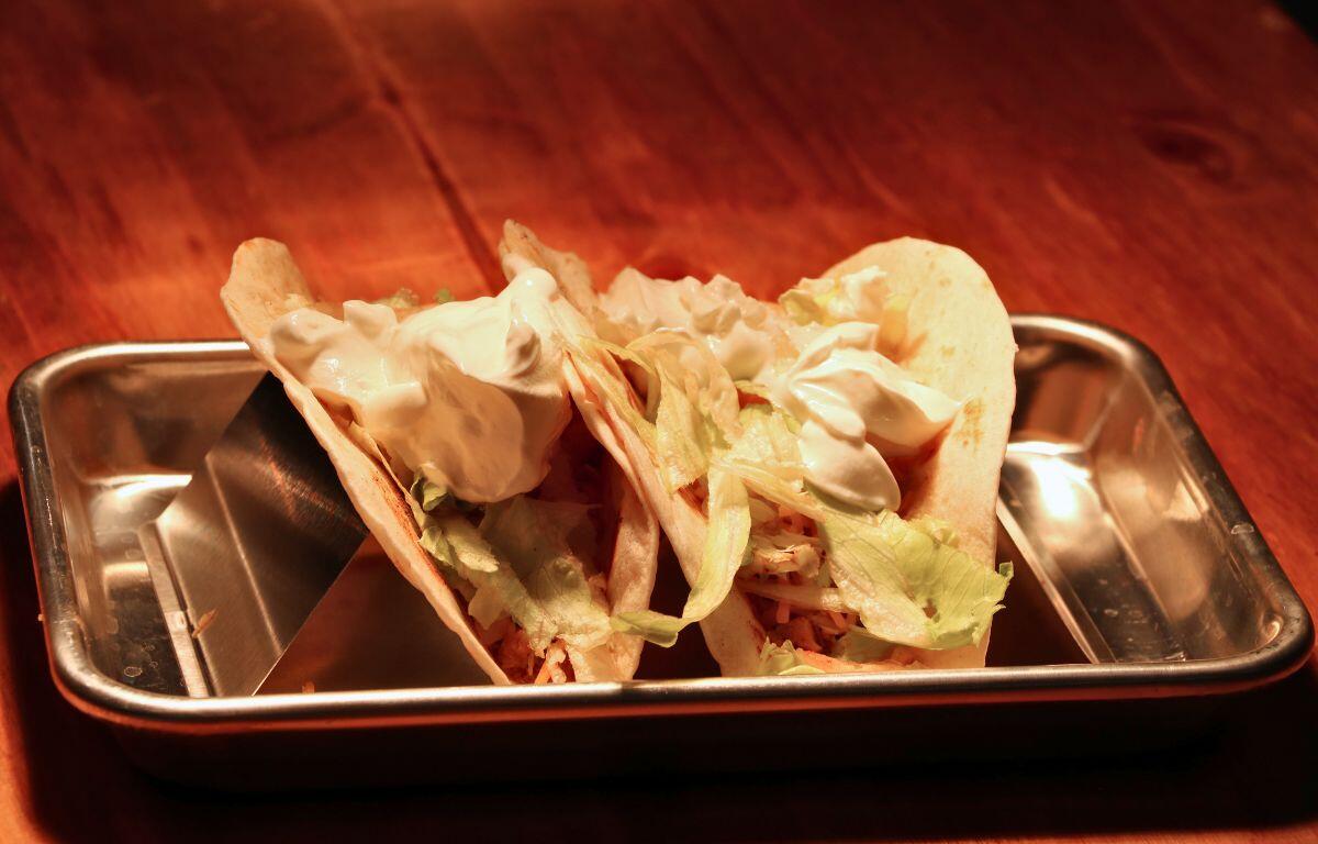 Two soft tacos filled with lettuce, shredded vegetables, and dollops of sour cream on a metal tray over a wooden table.