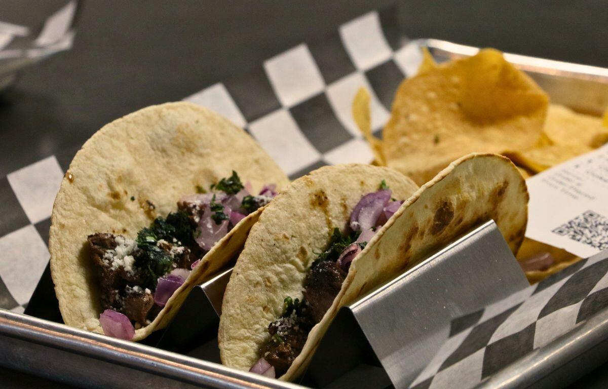 Two Tacos sit on a checkered paper on top of a metal tray with tortilla chips behind them.