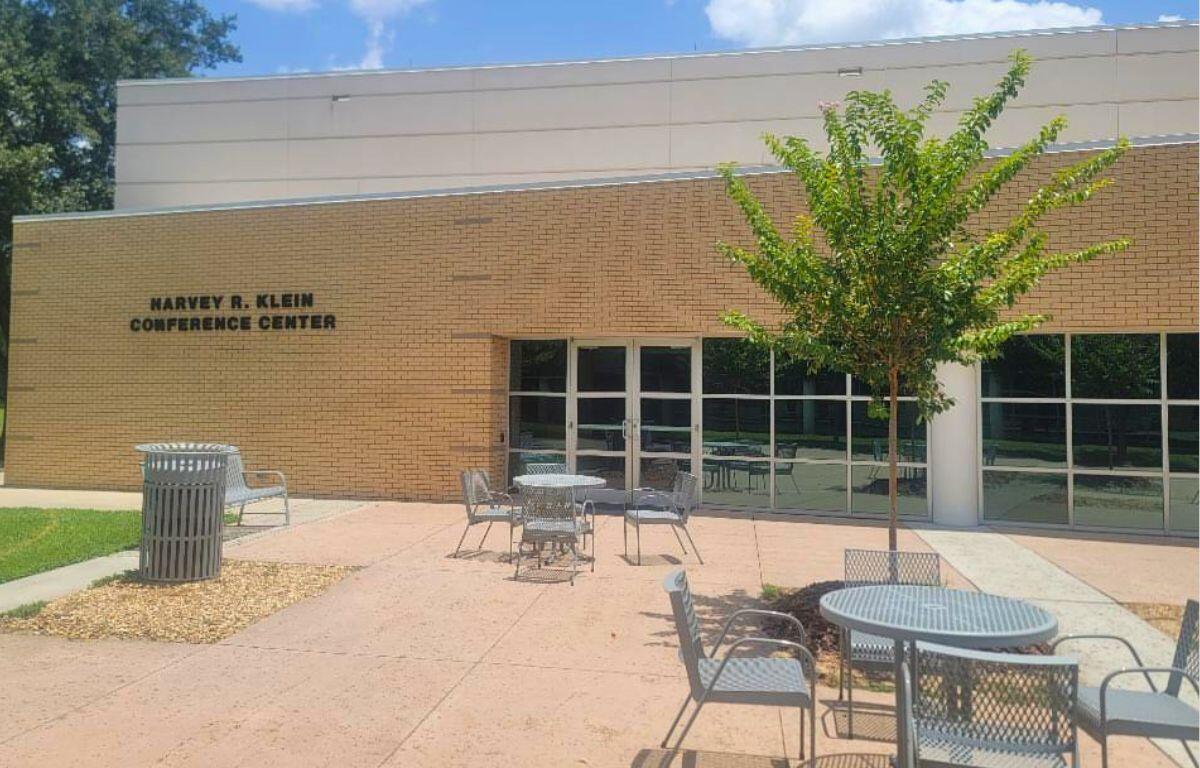 Brick exterior of Marvey R. Klein Conference Center with a tree, metal tables and chairs in a sunny courtyard.