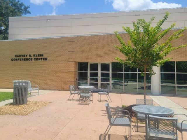 Brick exterior of Marvey R. Klein Conference Center with a tree, metal tables and chairs in a sunny courtyard.