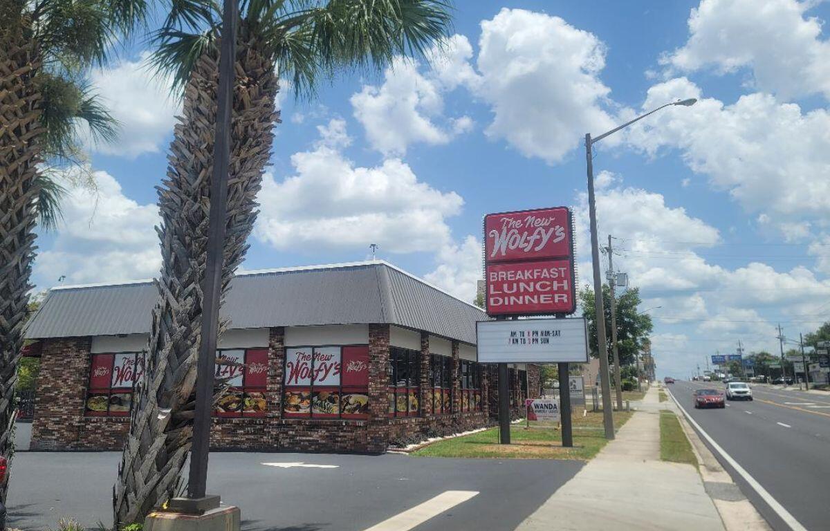 Restaurant exterior with brick walls and large red sign reading 'The New Wolfy's' near a sidewalk and palm trees.