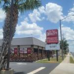 Restaurant exterior with brick walls and large red sign reading 'The New Wolfy's' near a sidewalk and palm trees.