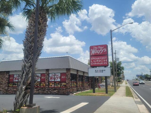 Restaurant exterior with brick walls and large red sign reading 'The New Wolfy's' near a sidewalk and palm trees.