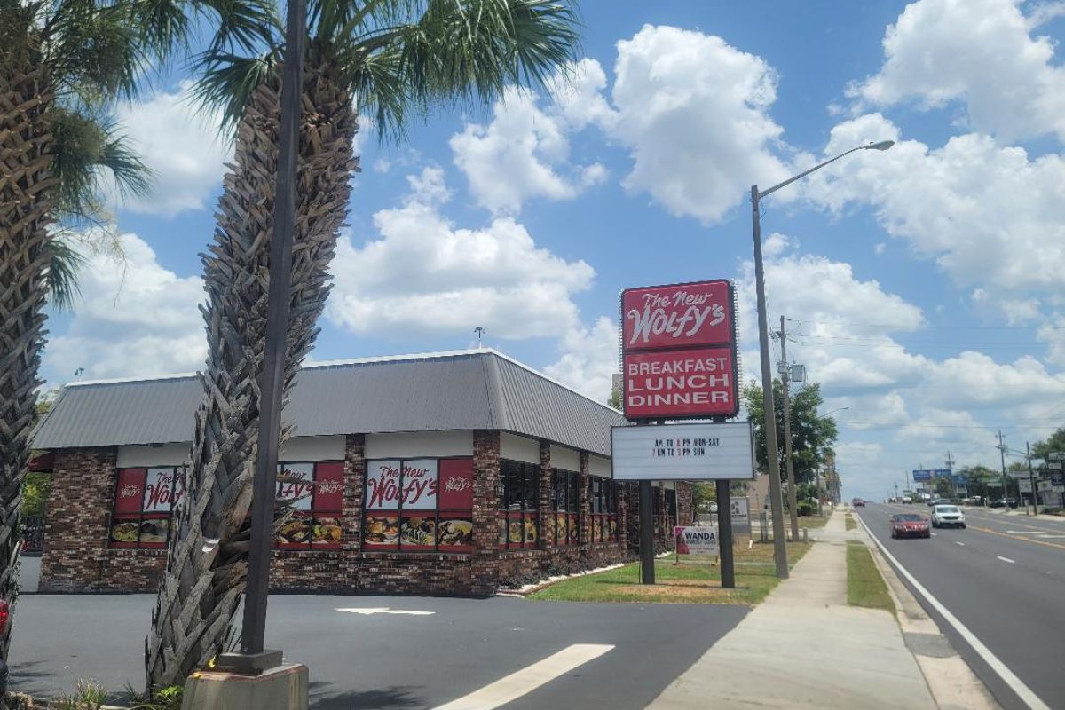 Restaurant exterior with brick walls and large red sign reading 'The New Wolfy's' near a sidewalk and palm trees.