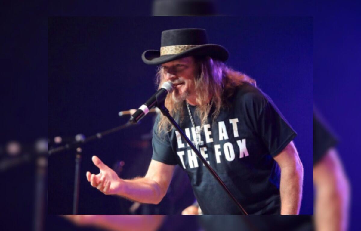 Male singer with long hair and a black hat performs into a microphone on a dimly lit stage; his shirt reads 'LIVE AT THE FOX'.