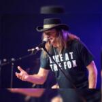 Male singer with long hair and a black hat performs into a microphone on a dimly lit stage; his shirt reads 'LIVE AT THE FOX'.