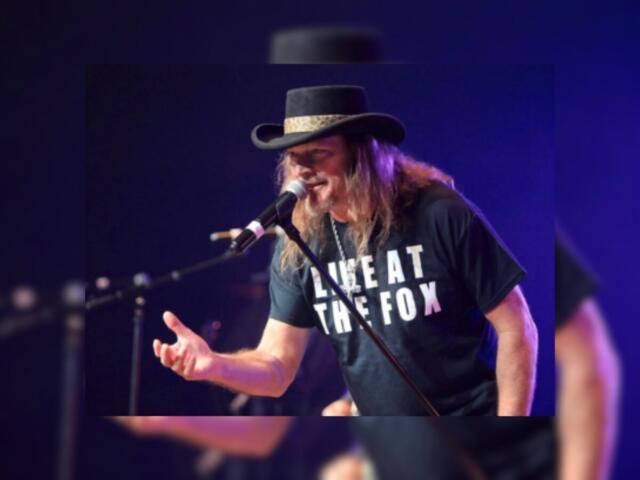 Male singer with long hair and a black hat performs into a microphone on a dimly lit stage; his shirt reads 'LIVE AT THE FOX'.