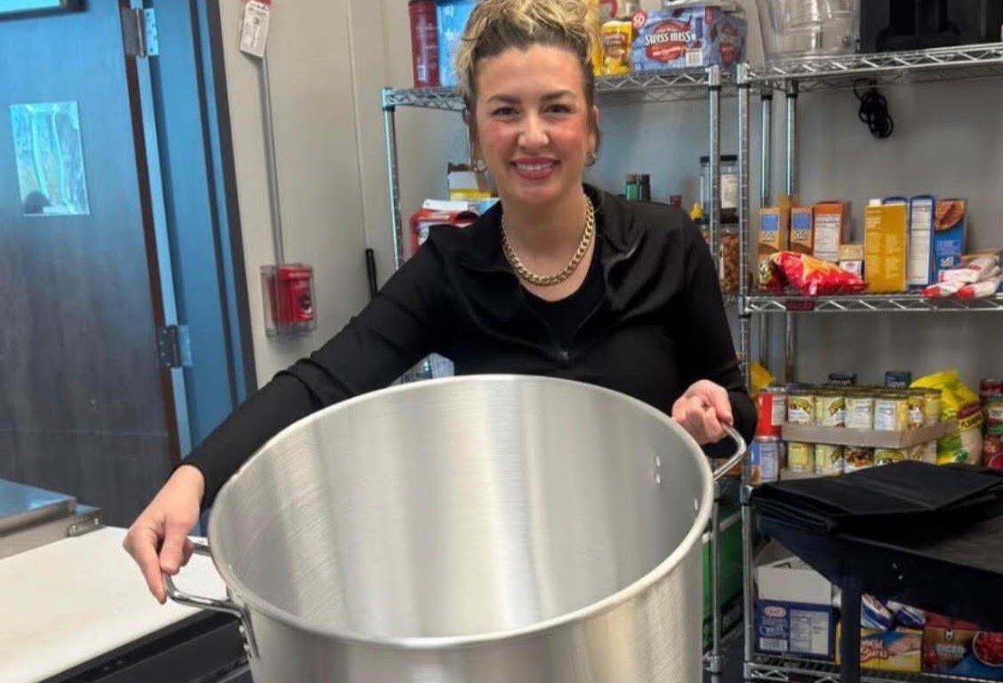 Smiling woman in a black outfit holds a large stainless steel pot in a busy kitchen with shelves of snacks and equipment in the background; a round Healthy Hub logo is in the lower left corner.
