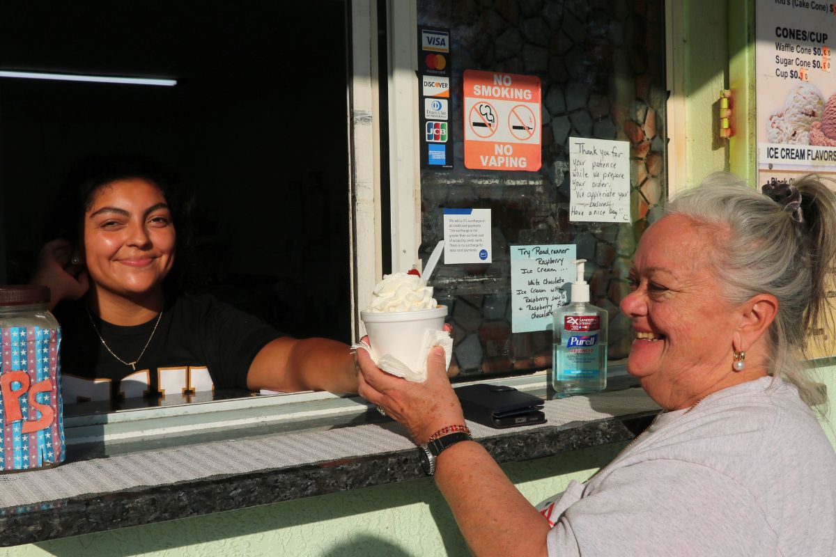 A restaurant window exterior. Woman hands another woman ice cream in a cup through a window.