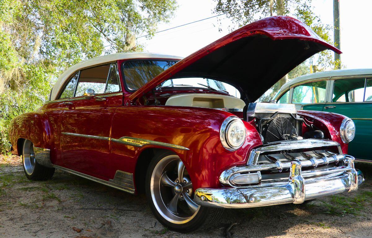 A vintage red car in a lot next to a wooded area.