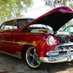 A vintage red car in a lot next to a wooded area.