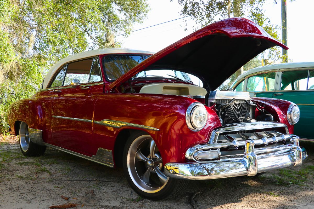 A vintage red car in a lot next to a wooded area.