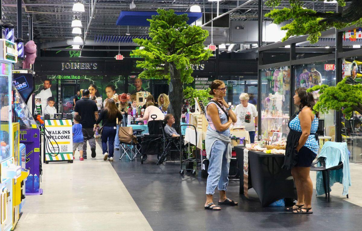 A crowd of guests and vendors walk through a variety of booths. Fake trees and arcade machines surround the area.