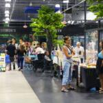 A crowd of guests and vendors walk through a variety of booths. Fake trees and arcade machines surround the area.
