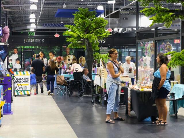 A crowd of guests and vendors walk through a variety of booths. Fake trees and arcade machines surround the area.