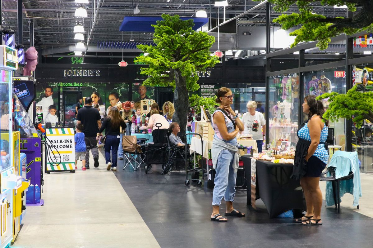 A crowd of guests and vendors walk through a variety of booths. Fake trees and arcade machines surround the area.