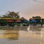 A town square with tables and umbrellas around an open area and fairy lights strung between supports.
