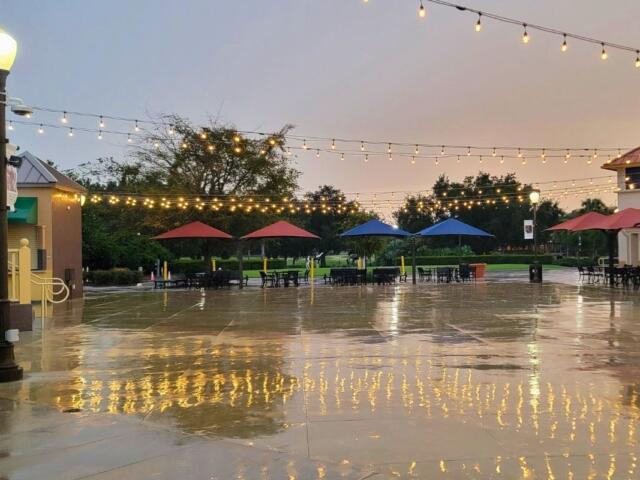 A town square with tables and umbrellas around an open area and fairy lights strung between supports.