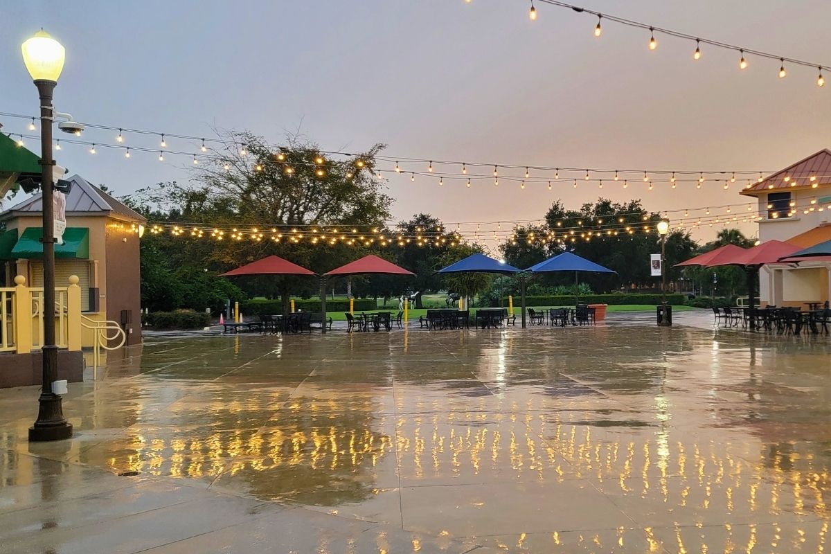 A town square with tables and umbrellas around an open area and fairy lights strung between supports.