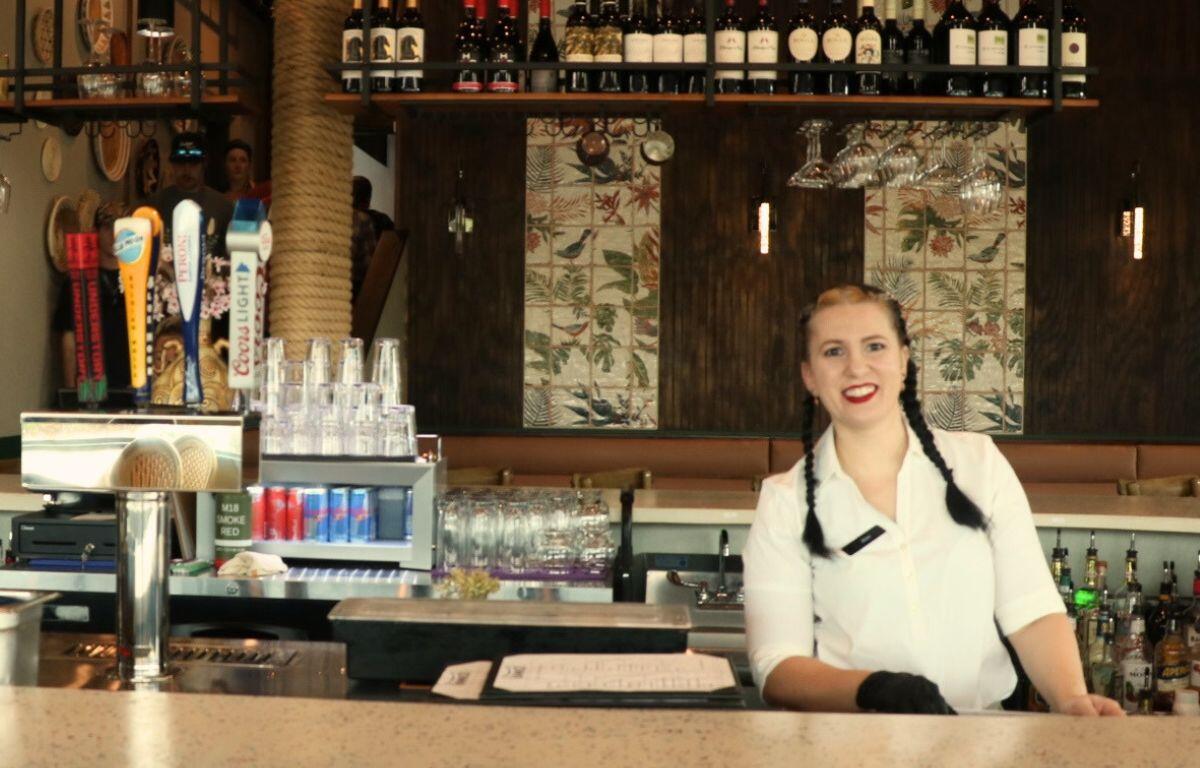 A woman stands behind counter at brightly lit bar.